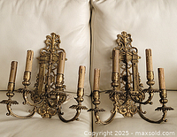 Pair of ornate brass wall sconces placed on a white leather surface showing their front with three candle-style arms each and detailed designs.