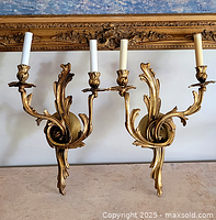 Pair of gold metal wall sconces with two candle-style arms each, ornate Rococo design, photographed with gold ornate frame in background
