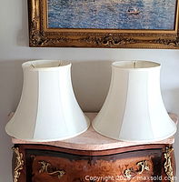 Two white fabric lamp shades sitting on a wooden surface with ornate brass decoration, front view.