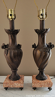 Pair of ornate metal table lamps with floral sculpted urn-style bases mounted on marble square bases with caster wheels, shown without lamp shades.