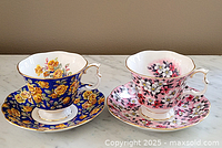 Two Royal Albert teacups and matching saucers, one in blue with yellow roses, one in pink with white and pink flowers, both with gold trim.