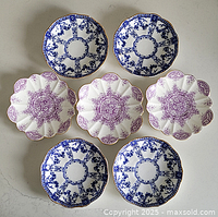 Seven porcelain saucers arranged showing four blue and white Royal Crown Derby saucers and three purple and white Wileman saucers with floral designs and scalloped edges.