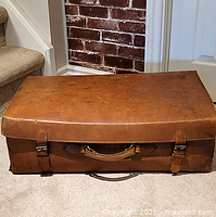 Front view of antique leather suitcase showing buckle straps and handle, placed on carpeted floor in front of brick wall and stairway.
