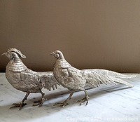 Pair of two silver-plated vintage bird figurines with detailed feather texture, standing on a marble surface against a brown background.