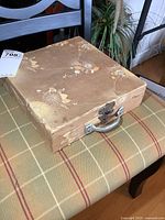 Closed vintage decorative carrying case with floral design, metal clasp, and handle, placed on a patterned chair.