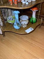 Three pieces of art glass on wooden shelf: two vases and one footed dish. Vases are about 9 inches tall with colorful designs; one vase is red/white/green, the other blue/white. The footed dish is amber and white with ruffled edges.