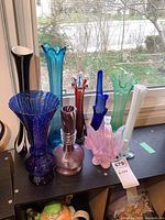 View of all nine art glass vases arranged on a shelf by a window showing variety of colors and shapes.