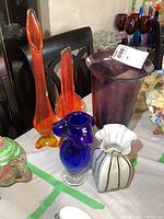 Six art glass pieces displayed on table (purple, two orange bud vases, blue, white-striped vase, orange swirl bowl)