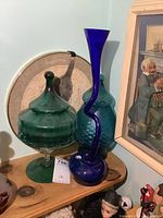 Green glass pedestal candy dish, tall cobalt blue twisted glass vase, and a blue glass canister shown on a wooden shelf against a pale blue wall.