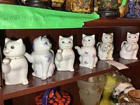 Shelf display of six small cat-shaped ceramic teapots in a row showing front views, different painted designs and colors on each.