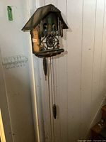 Full view of the cuckoo clock mounted on a wall, showing the clock body, roof, two weights hanging on chains, and overall color and wear.