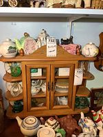 Small wooden display cabinet with two glass doors, antique style metal handles, and two small rounded shelves on right side. Cabinet contents shown but not included.