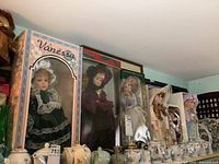 Six boxed porcelain dolls displayed on a shelf, dressed in period costumes with retail and collection boxes visible.