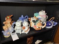 Full view of the shelf showing nine colorful ceramic teapots mostly cat-themed, including the large cat holding a fish, elephant, fish, and bunny shaped teapots.