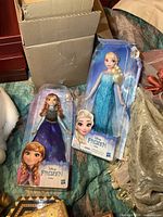 Photo showing both Elsa and Anna dolls in original packaging on a bed with a cardboard box behind them.