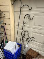 Seven metal garden shepherd hooks of varying sizes leaning against a garage door and wall, including the tallest approximately 7 feet in height