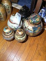 Photo shows five ginger jars/urns with lids of various sizes and decorative styles grouped on a wooden floor.