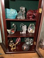 Wooden cabinet shelf with nine glass and resin animal figurines including rabbits, dogs, a fish, cats, and a gold-toned bird. Varied colors and translucency.