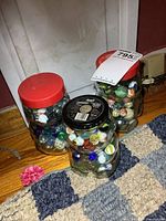 Photo of three glass jars filled with various vintage marbles including shooters. Jars are sealed with metal lids, placed on a wooden floor next to a carpet.