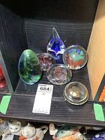 Shelf displaying six glass paperweights in various shapes and colors including teardrop, spherical, and flower designs inside clear glass.