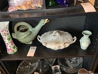 Display shelf with oval serving dish center, green pottery goose figurine on right, floral tall vase on left, and small green vase on far right.