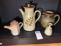 Four pottery items arranged on shelf: large speckled teapot with leaf design, geometric teapot, ceramic pitcher, and bud vase