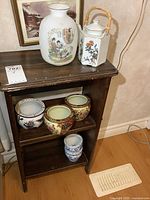 Photo showing large white porcelain vase with Asian figure paintings and white hexagonal teapot with bamboo handle on top shelf of wooden shelf unit, plus five smaller round planters on lower shelves.
