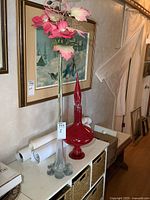 Both clear vase with faux flowers and red decanter displayed on table under framed art