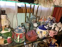 Wide view of medium to large sized ceramic teapots of various styles including leaf and berry design, blue and white, figurative house, and snowman set on glass shelf.