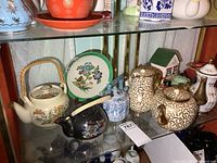 Shelf of assorted teapots including black floral teapot, floral cream-colored teapot, blue and white oriental vase, decorative floral plate, and small decorative house with green roof.
