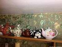 Shelf holding eight assorted ceramic teapots with various designs and colors against green leafy wallpaper background