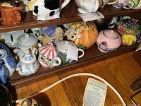 Bottom shelf of cabinet containing nine various ceramic teapots in different shapes and colors