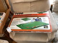 Top view of the Tyco Track Layout Expander Kit box sitting on a beige chair showing brand and product name with graphics of train layout.