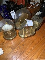 Four vintage clocks on wooden floor: three glass dome anniversary clocks with visible brass mechanisms and one old brass wind-up clock with square shape.