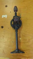 Antique metal hand drill standing on a wooden surface, showing front and base details.