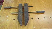 Antique wood clamp resting horizontally on a wooden workbench, showing the front and top of the clamp, wooden jaws, and metal rods with wooden handles.