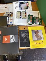 Overview of art papers, sketch pads including Canson and Bristol boards, water-soluble oil pastels, inks, and brushes arranged on table.