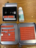 Photo showing black-handled stamp applicator in metal tin, bottle of Making Memories rubber stamp cleaner, and two silver tins containing red rubber alphabet and number stamps.