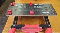 Top view of black rectangular work surface with printed measurement grid, Craftsman logos, and four red clamps.