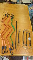 Photo showing nine rubber bungee cords in various lengths and colors (black, orange, green) laid out on a wooden surface with metal S and J hooks on ends.