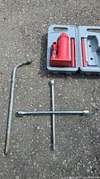 Photo showing the cross wrench, lug wrench, and hydraulic jack in open carrying case on asphalt surface.