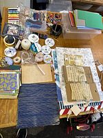 Overview of assorted crafting supplies including ribbons, embroidery floss, hemp cords, thread, and paper materials arranged on a table with a clear plastic storage bin.