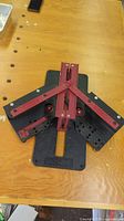 Top-down view of Magic Mitre tool laid flat on wooden surface showing red and black plastic components, pivoting arms, and slots for angle adjustment.
