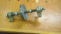Metal pulley assembly placed on wooden surface, showing pulley wheel, shaft, and bearing housings from an angle.