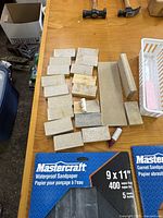 Photo showing hand-sanded wooden blocks, two packs of Mastercraft sandpaper sheets (400 grit and 320 grit), and small container with craft items.