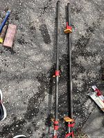 Two 48 inch pipe clamps with black metal bars, red metal clamping mechanisms, and yellow plastic pads, lying on asphalt surface.