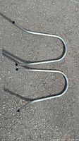 Two silver steel ladder stand-off arms placed on pavement, showing the curved profile and hollow tube construction.