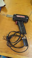 Black Weller Expert soldering iron tool with pistol grip, red trigger, soldering tip, and attached power cord lying on wooden surface