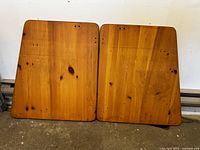 Two wood table tops placed side by side on floor against wall showing full top surfaces with visible wood grain and knots.