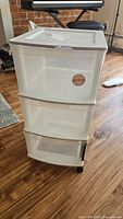 Front view of the 3-drawer plastic storage unit with white frame and clear drawers, showing sticker on top drawer and caster wheels at base.